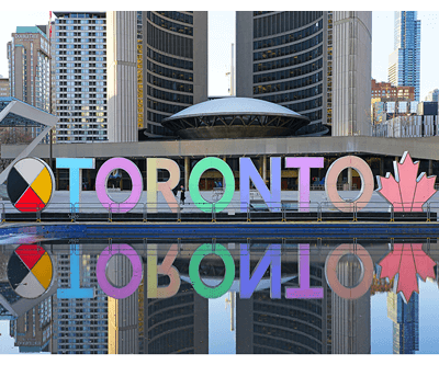 Large, colorful letters spelling “TORONTO” with a medicine wheel symbol and pink maple leaf, reflected in a pool of water at Nathan Phillips Square, with modern buildings in the background.