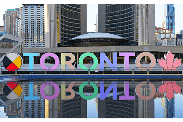 Large colorful “TORONTO” sign with a medicine wheel on the left and a maple leaf on the right, reflected in water, set in front of modern high-rise buildings at Nathan Phillips Square.