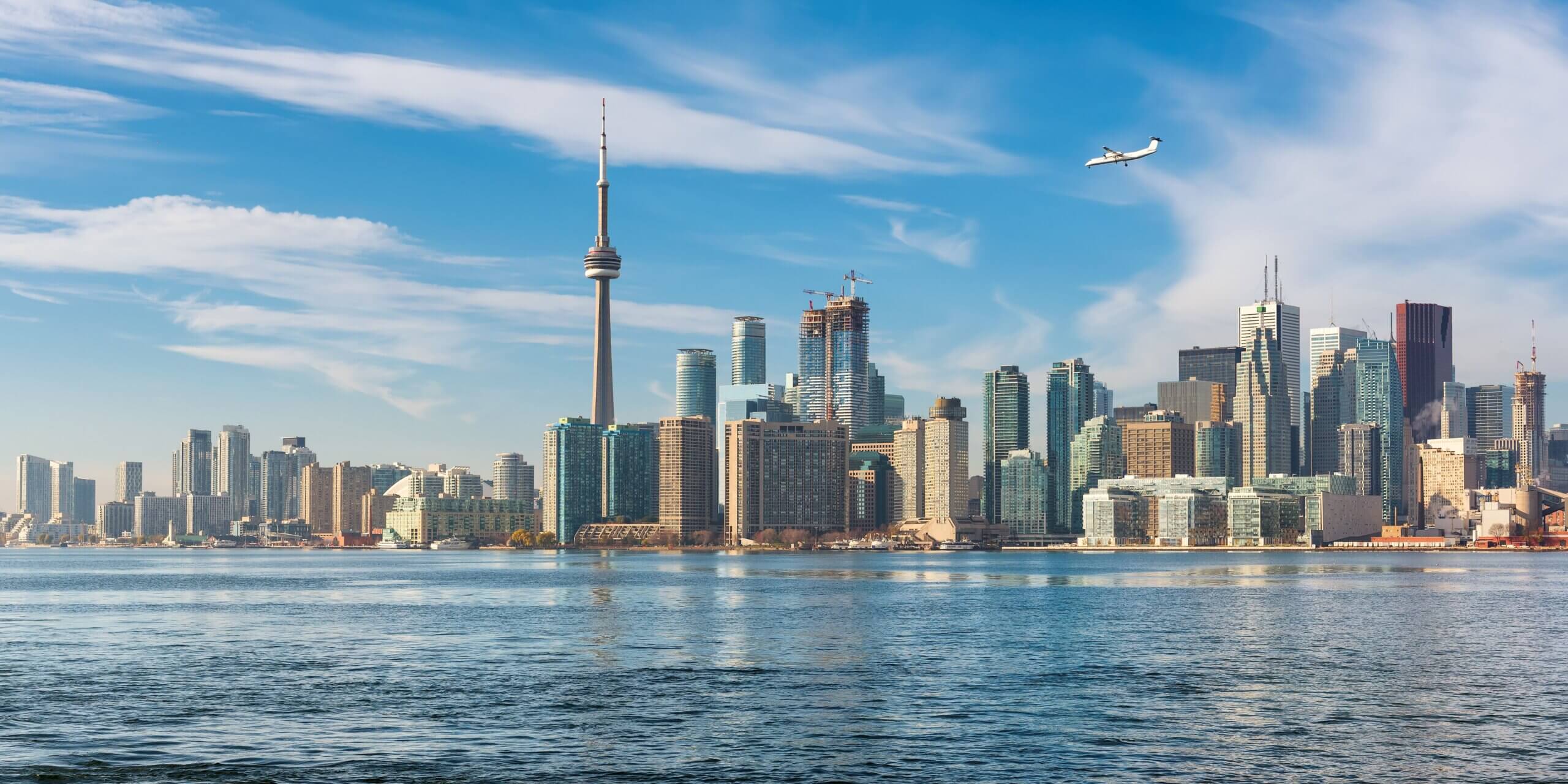 Toronto skyline with tall buildings, including the CN Tower, under a blue sky with wispy clouds. A small airplane flies above the city. The foreground shows calm water reflecting the skyline.