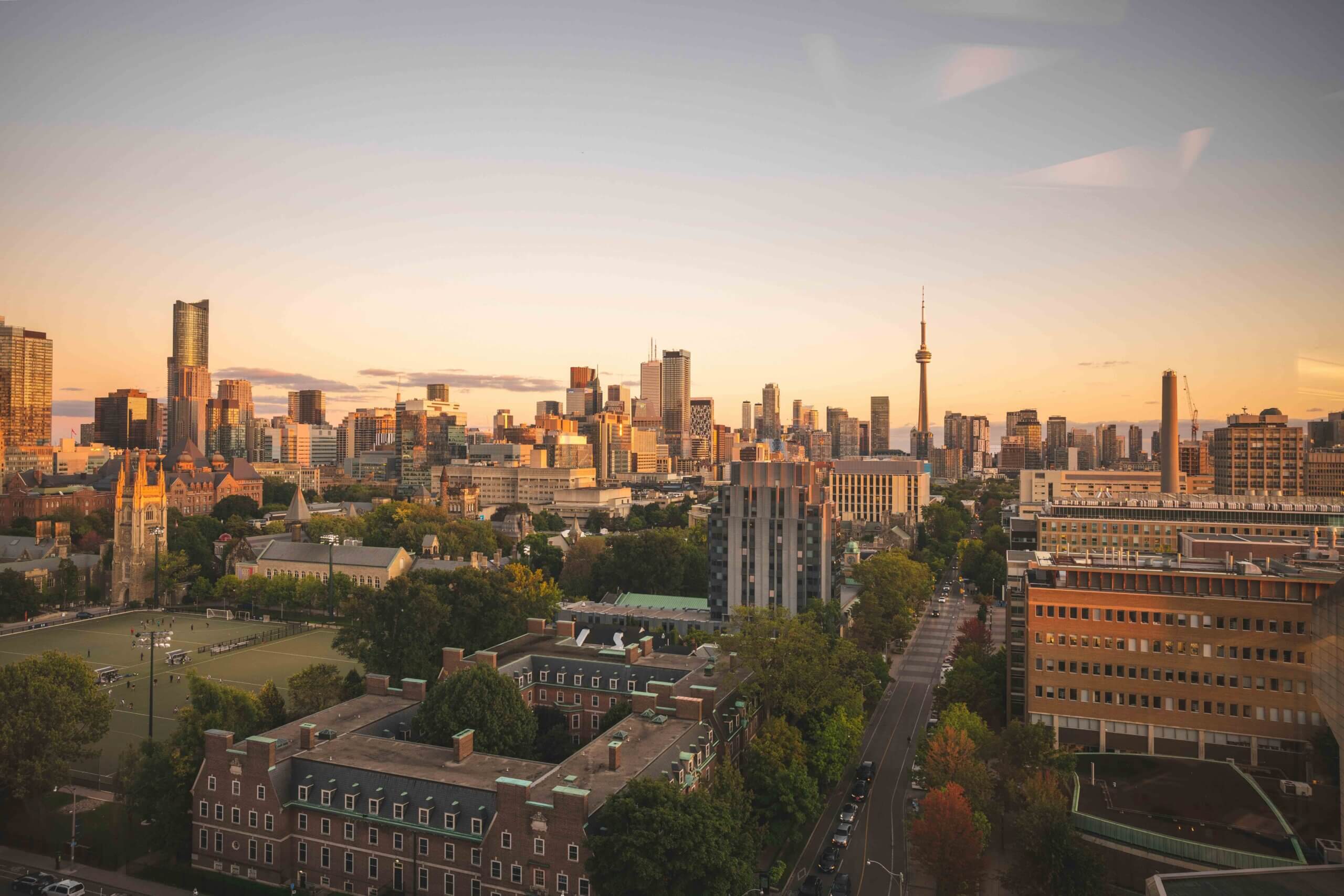 A wide view of Toronto’s skyline at sunset, featuring the CN Tower and modern high-rise buildings, with trees, historic buildings, and a sports field in the foreground under a clear sky.
