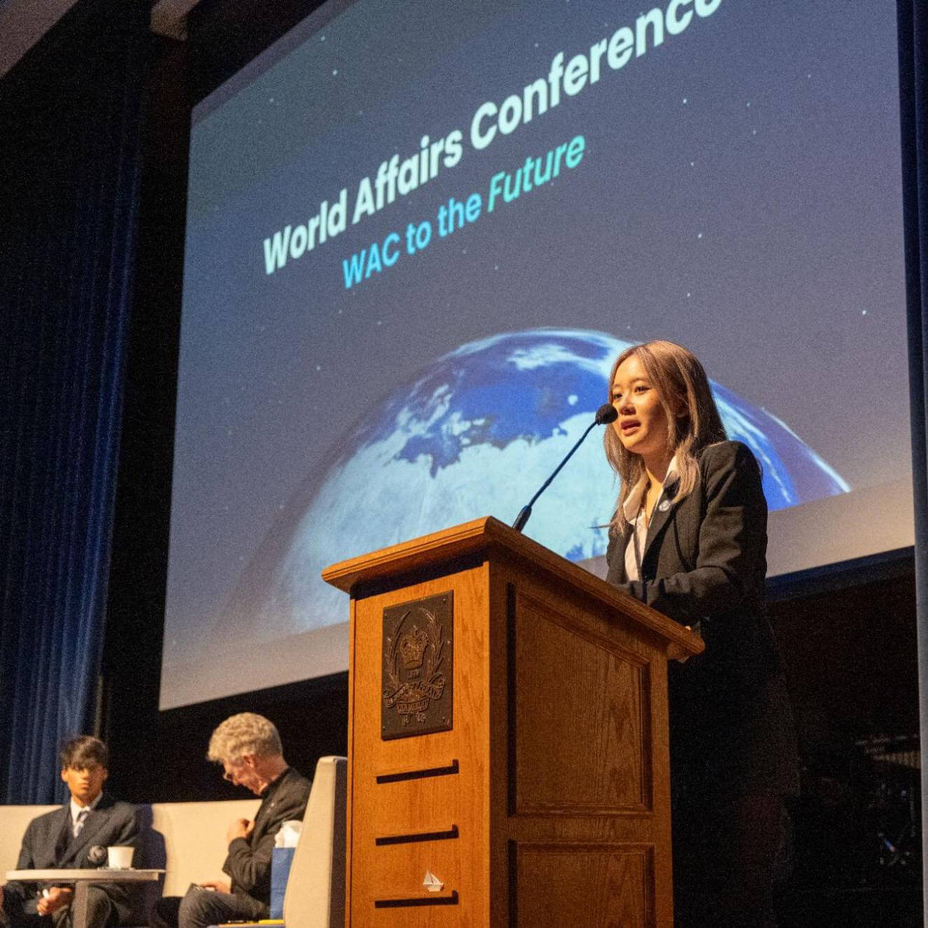 A woman in a suit speaks at a podium on stage during the World Affairs Conference, with two men seated behind her and a large screen displaying an image of Earth and conference details in the background.
