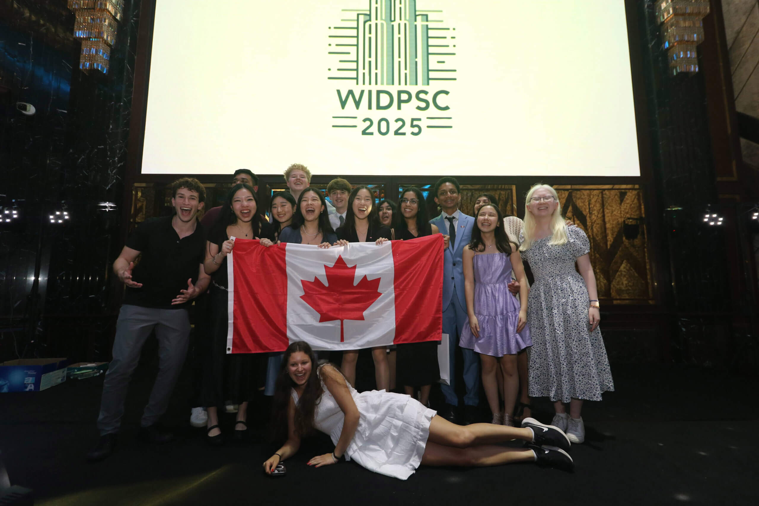 A group of smiling young people pose on stage with a Canadian flag in front of a large screen displaying "WIDPSC 2025." One person lies playfully in front of the group, all appearing celebratory and excited.