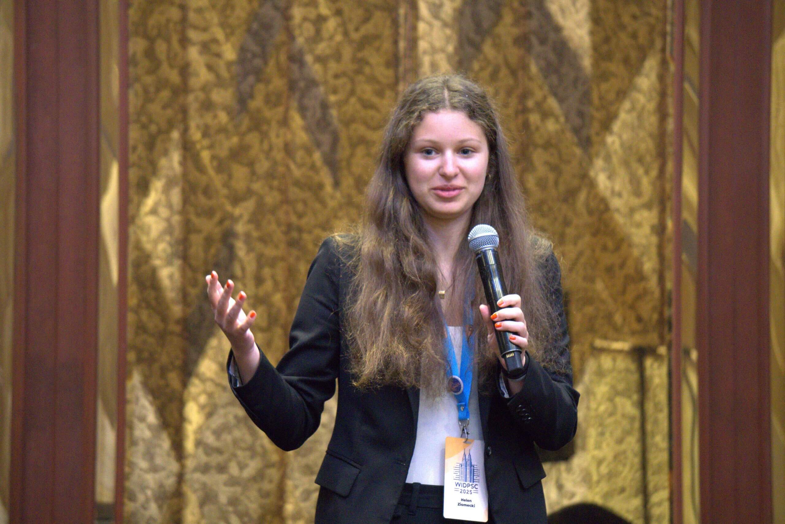 A young woman with long brown hair, wearing a black blazer and name badge, holds a microphone and gestures while speaking in front of a gold-patterned curtain.