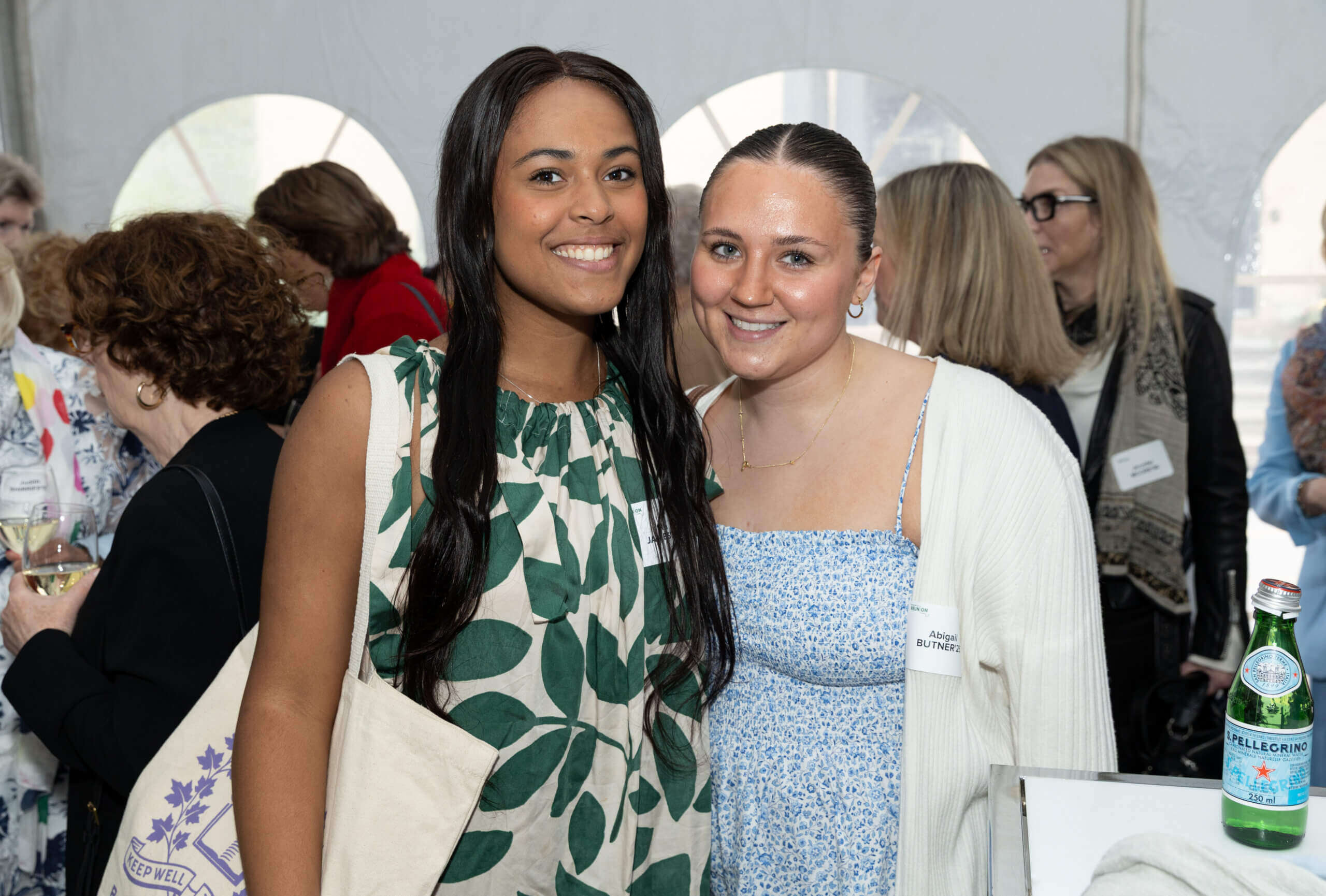 Two women smiling and posing together at an indoor social event. One wears a green leaf-patterned dress, the other a blue patterned dress with a white sweater. People mingle in the background, and a bottle of sparkling water is on the table.