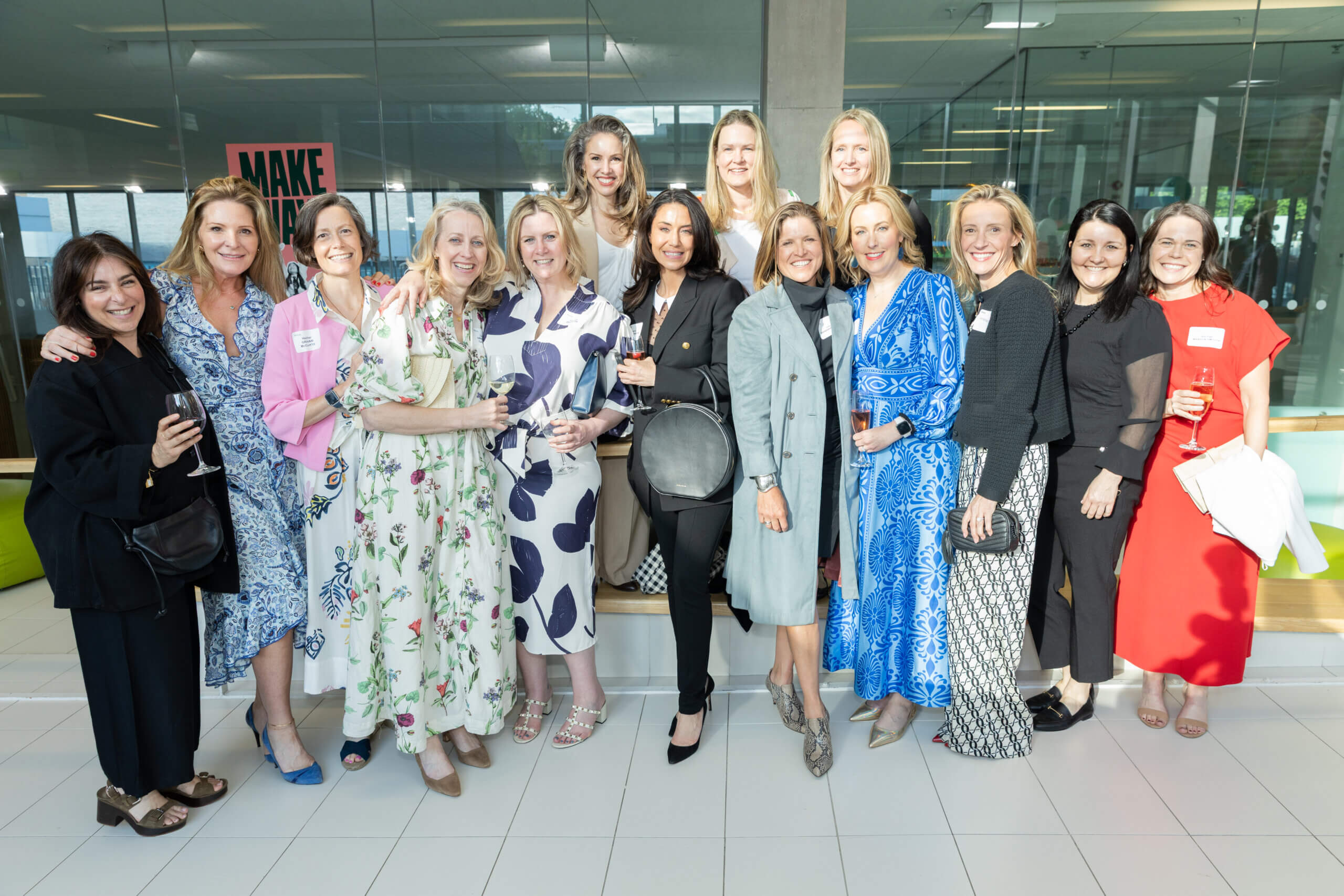A group of fourteen women stand together indoors, smiling at the camera. Most are dressed in semi-formal, colorful outfits and some hold drinks. The background features large windows and a sign that reads "MAKE.