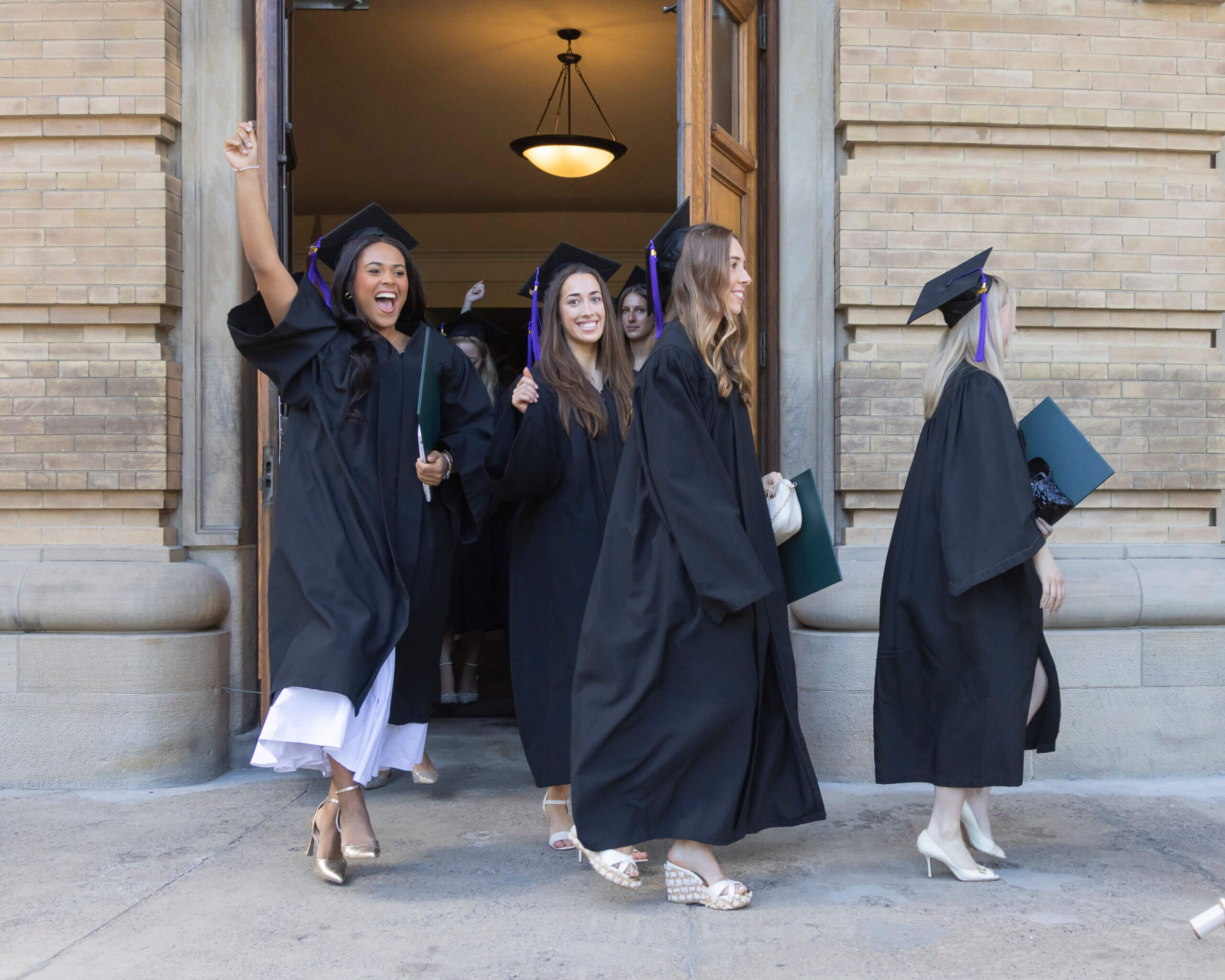 A group of graduates in black gowns and caps joyfully exit a building, smiling and celebrating. One woman raises her arm in excitement, while others walk beside her, holding diplomas.