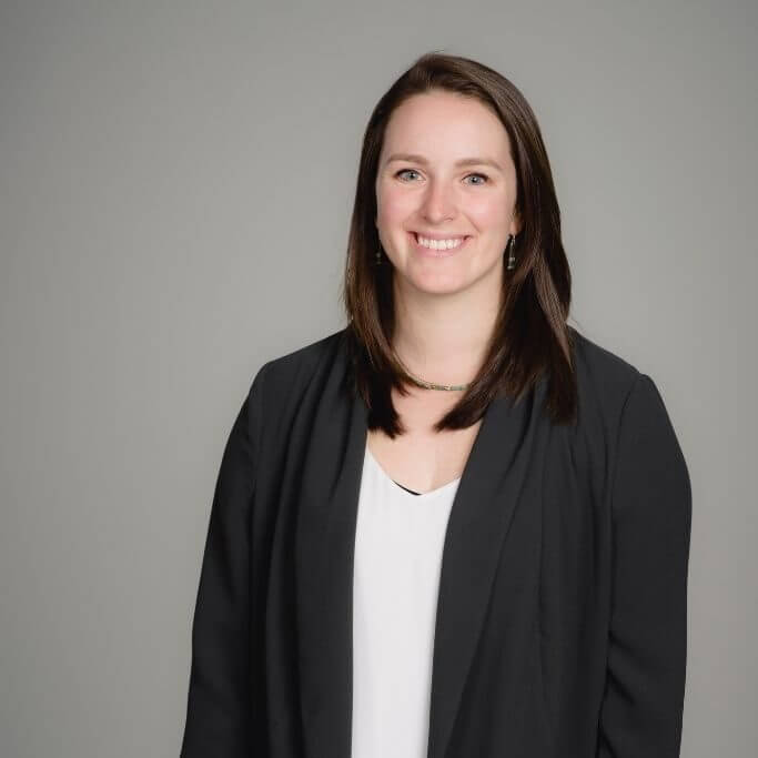 A woman with straight brown hair, wearing a black blazer over a white top, smiles at the camera against a plain, light gray background.