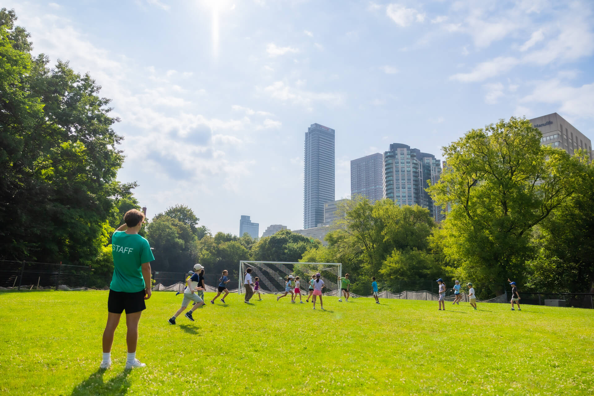 A group of children play soccer on a grassy field in a city park, with a staff member watching nearby. Tall buildings and green trees are visible in the background under a partly cloudy sky.