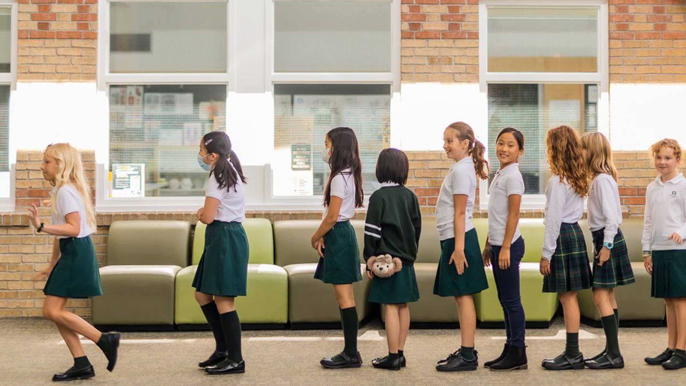 A group of young girls in school uniforms stand in a line indoors, some facing forward and some turning to the side. They are in front of a brick wall with windows and green couches.