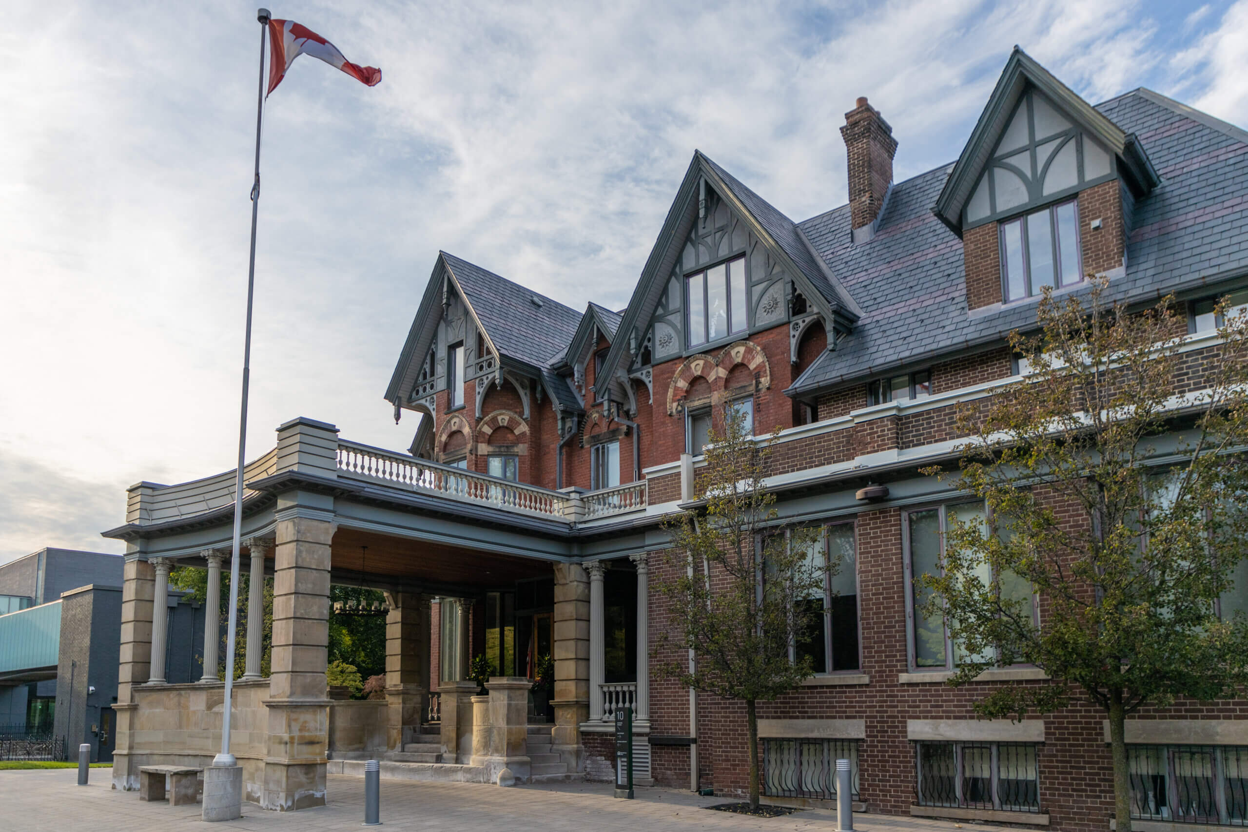 A large, historic brick building with peaked roofs, detailed architecture, and a covered entrance stands beneath a flagpole flying a red and white flag. Trees and a paved walkway are in the foreground.