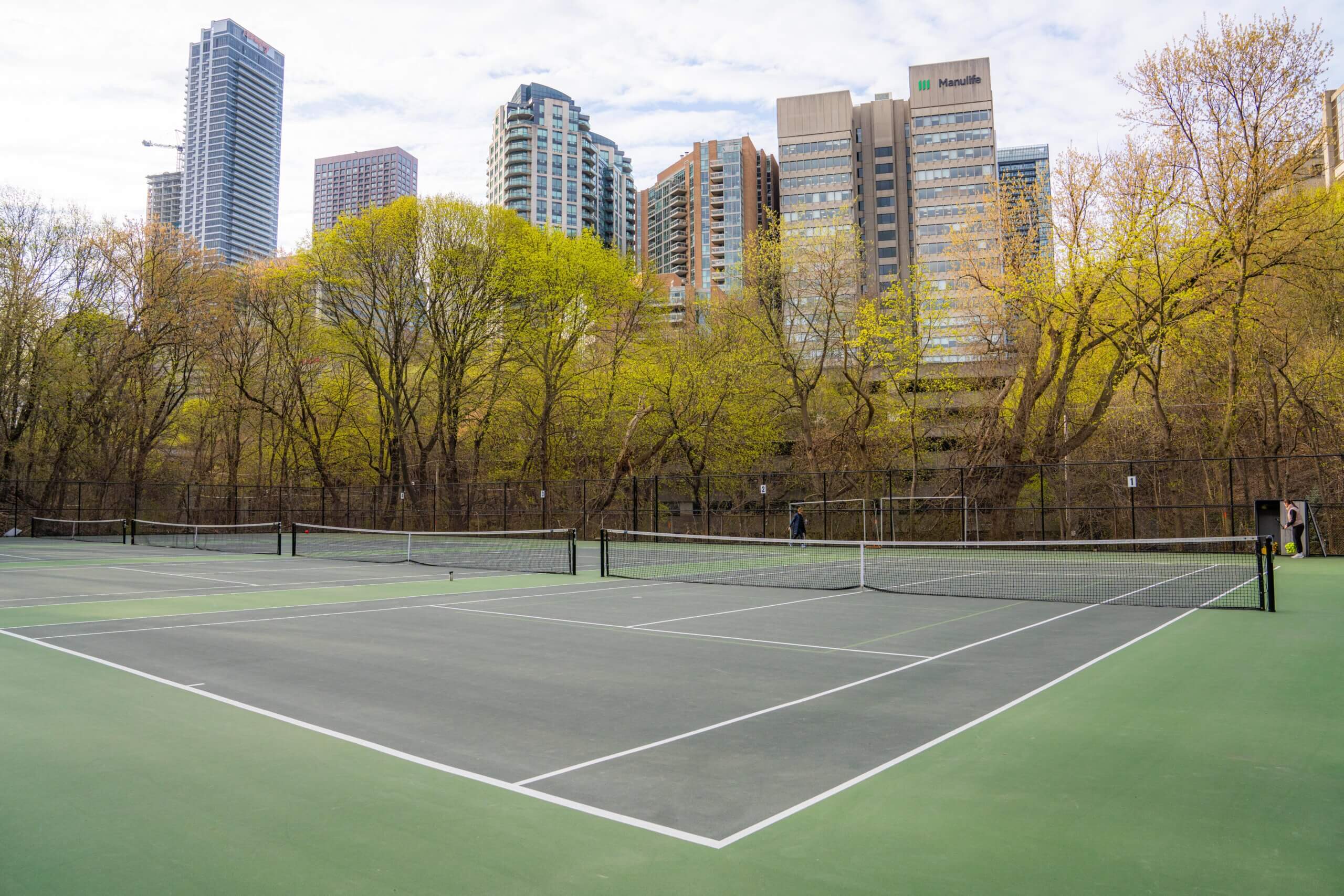 An empty outdoor tennis court with fresh markings, surrounded by trees with spring foliage, and tall city buildings in the background under a partly cloudy sky.