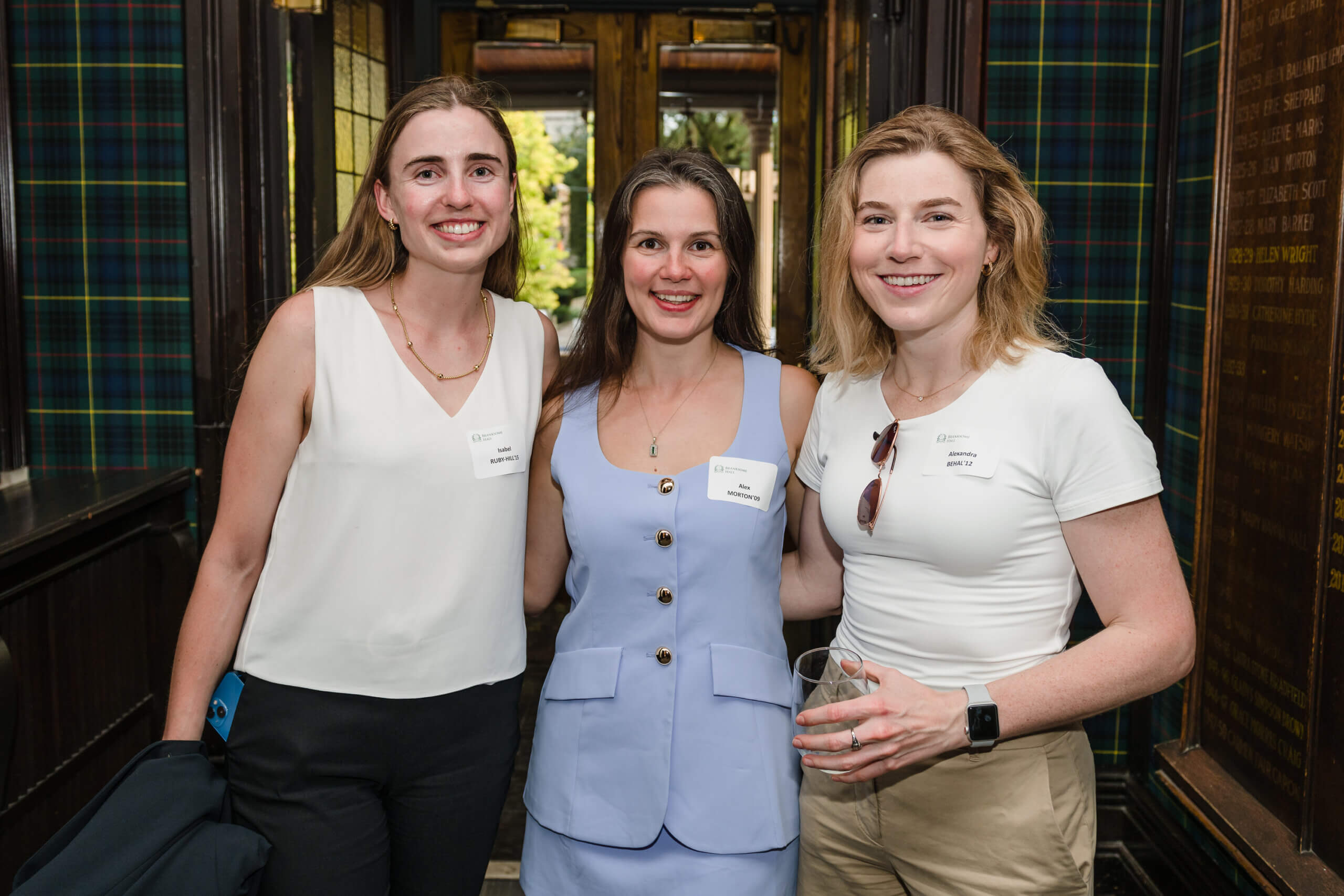 Three women stand close together and smile at the camera indoors. Two wear white tops and one wears a light blue vest and skirt. They have name tags, and one woman holds a drink. The background features dark wood and plaid walls.