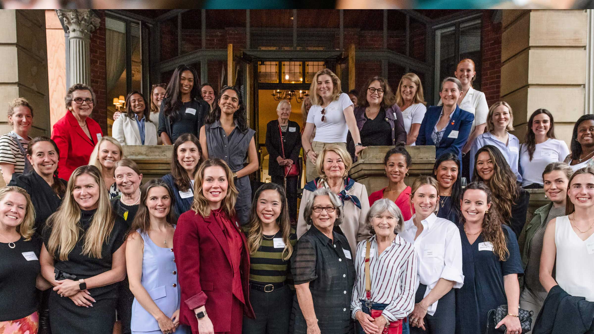 A large group of women, diverse in age and ethnicity, are smiling and posing together on the steps of a building, some standing and some sitting, dressed in business and casual attire.