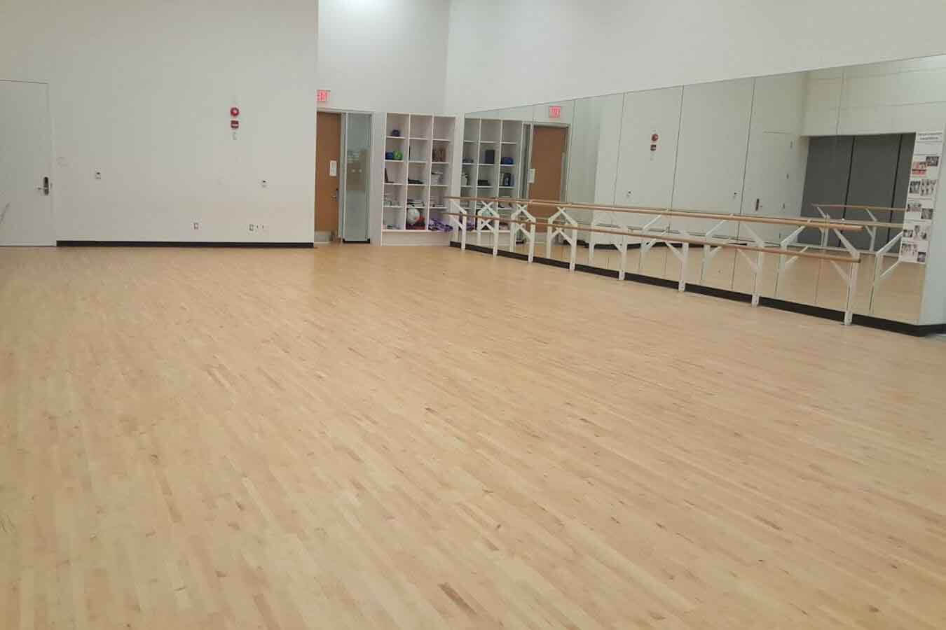 A spacious, empty dance studio with light wooden floors, mirrored walls, ballet barres, and shelves with various equipment against the back wall. Bright overhead lighting illuminates the room.