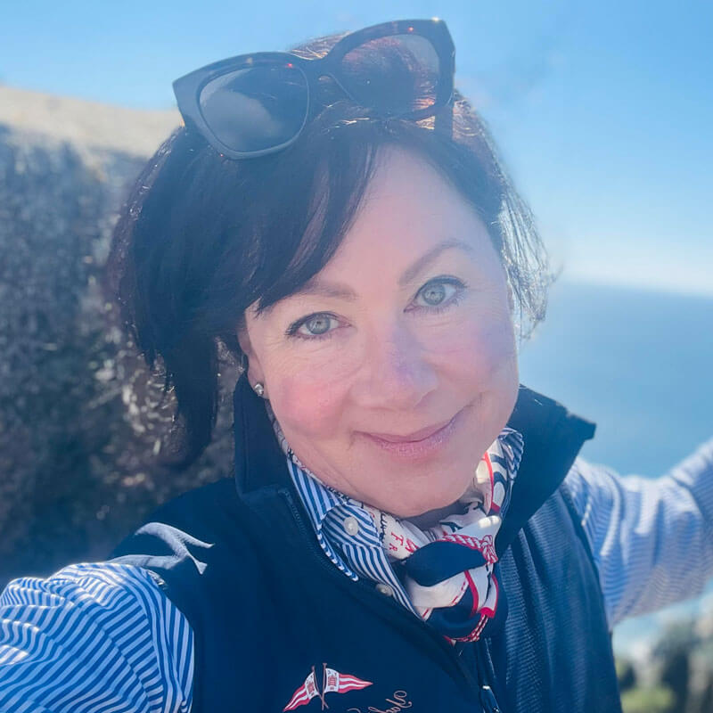 A woman with short dark hair and green eyes smiles outdoors, wearing sunglasses on her head, a striped shirt, a navy vest, and a patterned scarf. The background is bright and appears to show a sunny, coastal landscape.