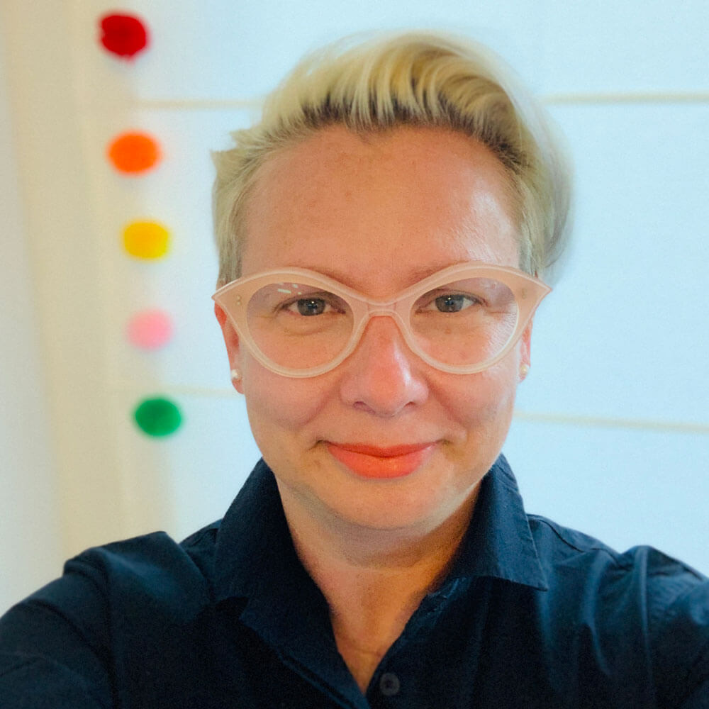 A person with short blond hair and light pink cat-eye glasses smiles at the camera. They wear a dark collared shirt. Six colored dots are arranged vertically on the white wall behind them.