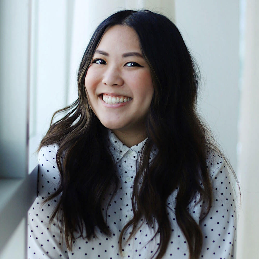 A woman with long, wavy dark hair smiles while wearing a white button-up shirt with black polka dots. She is standing indoors near a window with soft natural light.