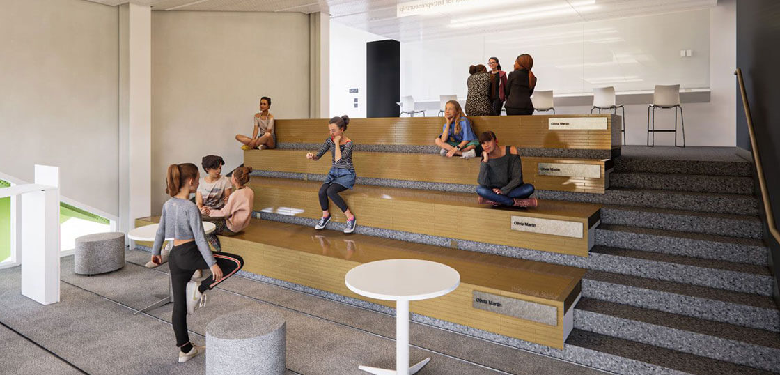 Several people sit and talk on tiered wooden seating in a modern, open indoor space with a whiteboard, tables, and stools. The atmosphere is casual and collaborative.