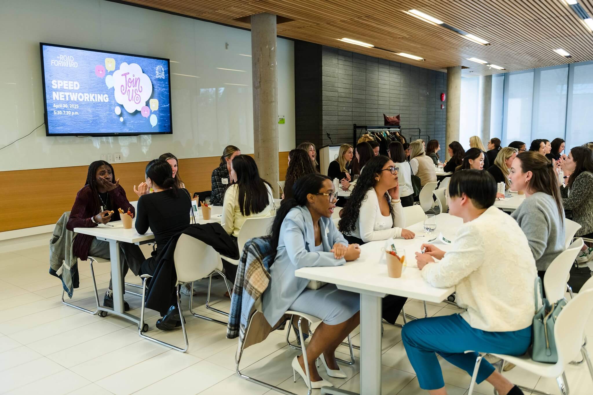A group of women sit at tables in a modern, bright room participating in a speed networking event. A large screen on the wall displays event details. The atmosphere is lively and professional.