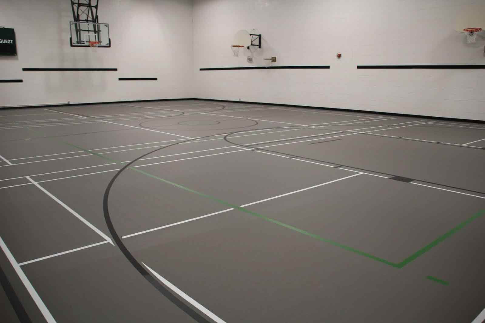 An empty indoor gymnasium with gray flooring featuring multiple white and black court lines, basketball hoops mounted on white walls, and no people present.