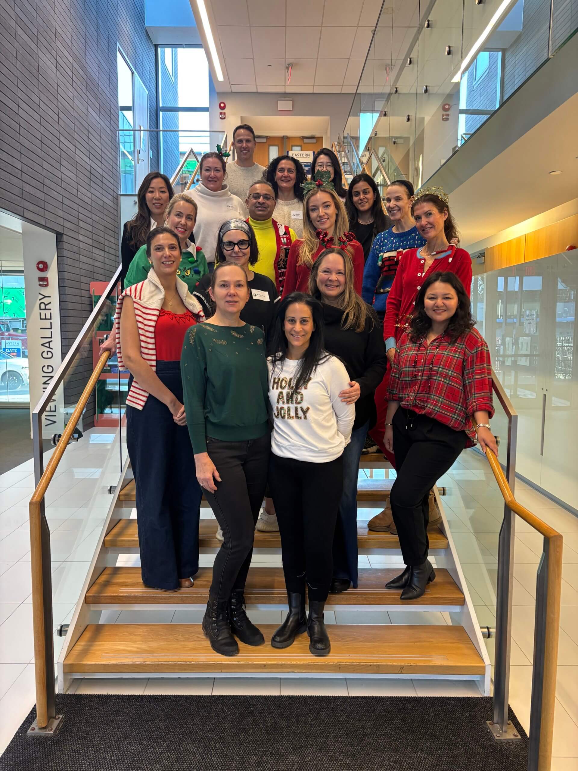 A group of 18 adults stand on a staircase indoors, smiling at the camera. Most are wearing festive or casual clothing, and natural light comes in through large windows in the background.