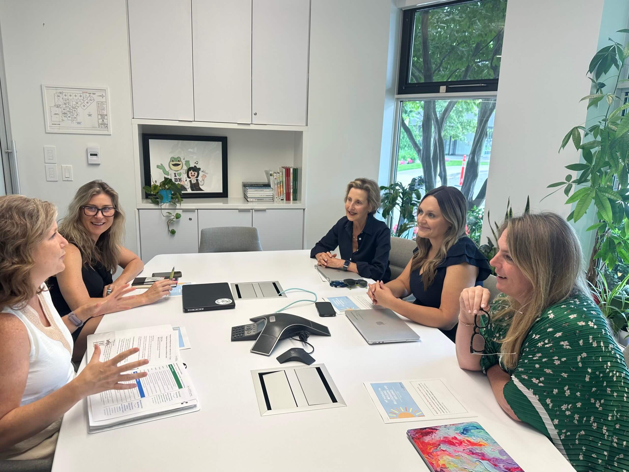 Five women sit around a conference table in a bright office, engaged in discussion. Laptops, documents, and a phone speaker are on the table. Large windows and plants surround the group, creating an open, collaborative atmosphere.
