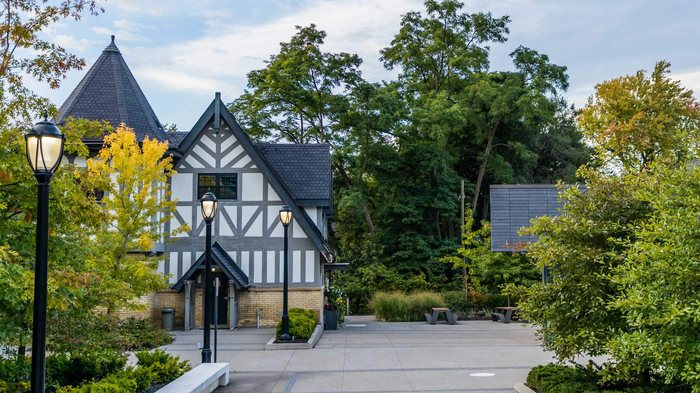 A Tudor-style building with exposed wooden beams and steep roof sits beside a tree-filled courtyard with benches and lampposts on a sunny day.