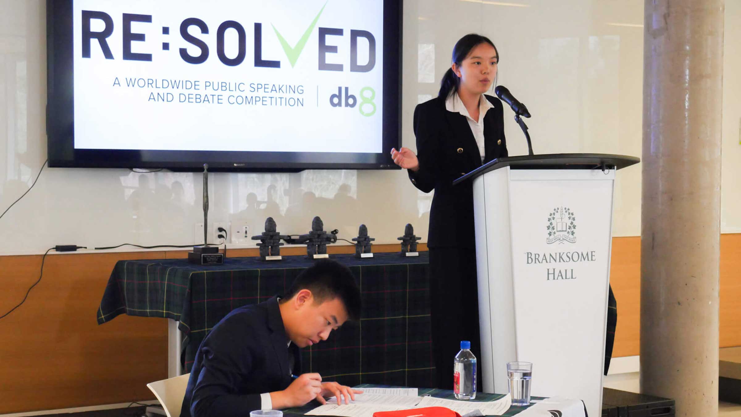 A young woman speaks at a podium labeled "Branksome Hall" during a debate competition, while a young man sits and writes at a table. A large screen behind them displays "RE:SOLVED" and competition details.