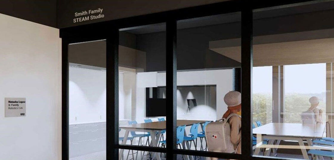 A person wearing a light coat and hat stands outside a glass-walled room labeled "Smith Family STEAM Studio," looking inside at tables, blue chairs, and large windows with a view.