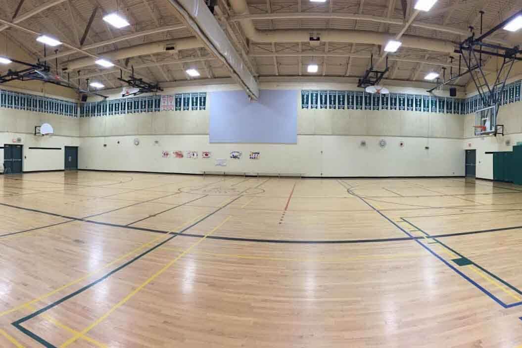 Wide view of an empty indoor gymnasium with a wooden floor, basketball hoops, high ceiling with lights, and banners hanging along the upper walls. The space is well-lit and has various court markings.