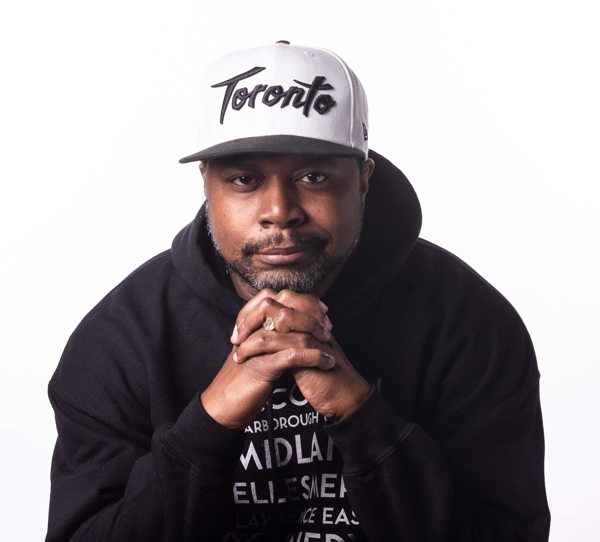 A man wearing a gray "Toronto" cap and a black hoodie sits with his hands clasped in front of his face, looking directly at the camera against a plain white background.