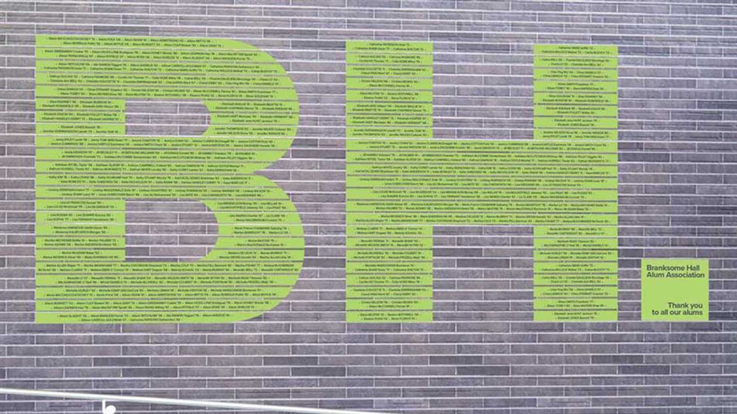 Large green letters "BH," made of many small names, are displayed on a gray brick wall. A yellow box at the right reads: “Bancone Hall Alumni Association. Thank you to all our alums.”.