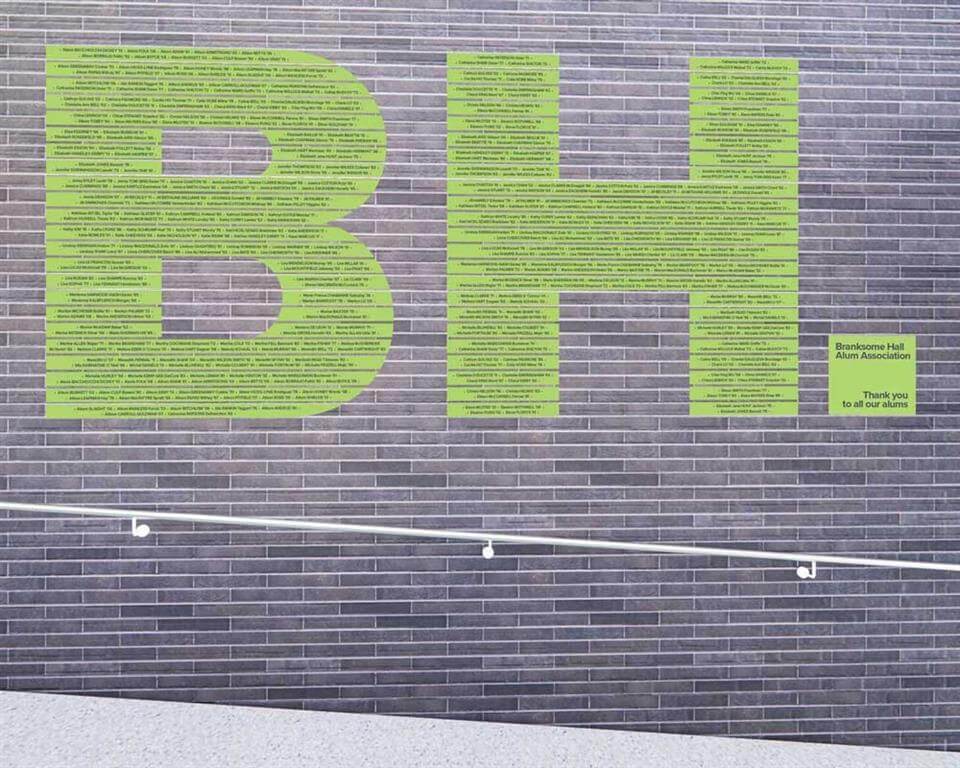 Large green letters "BH" made up of many small names cover a gray brick wall. To the right, a small green sign reads, "Bronfman Hall Alumni Association. Thank you to all our alumni." A white handrail runs along the wall.