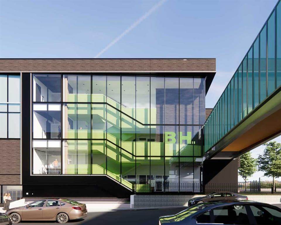 A modern building with large glass windows reveals green interior staircases. The letters "BH" are visible inside. A glass walkway connects to another building. Several cars are parked in front.