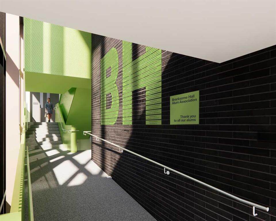 A modern hallway with dark brick walls and large green "BH" letters, featuring a handrail and a green sign thanking alums. Sunlight streams in, and a person is visible in the distance near the stairway.