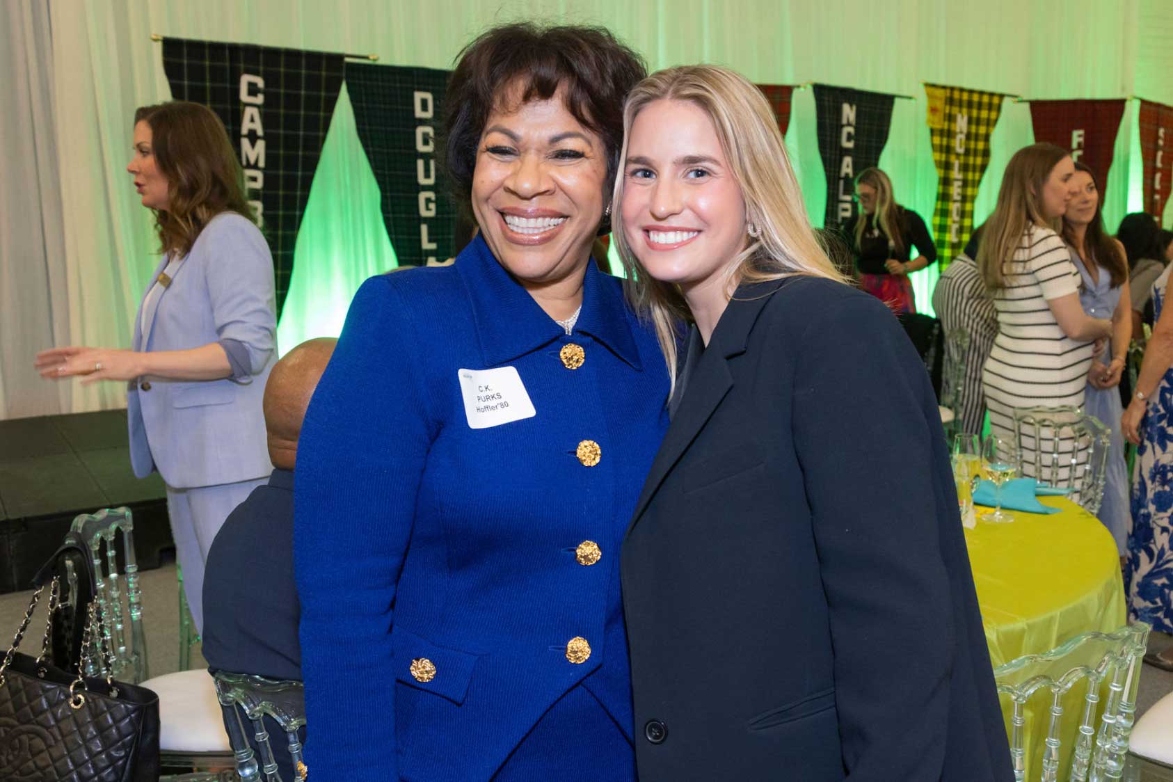 Two women smile and pose together at an indoor event. The woman on the left wears a royal blue blazer with gold buttons and a name tag, while the woman on the right wears a black blazer. Colorful banners hang in the background.