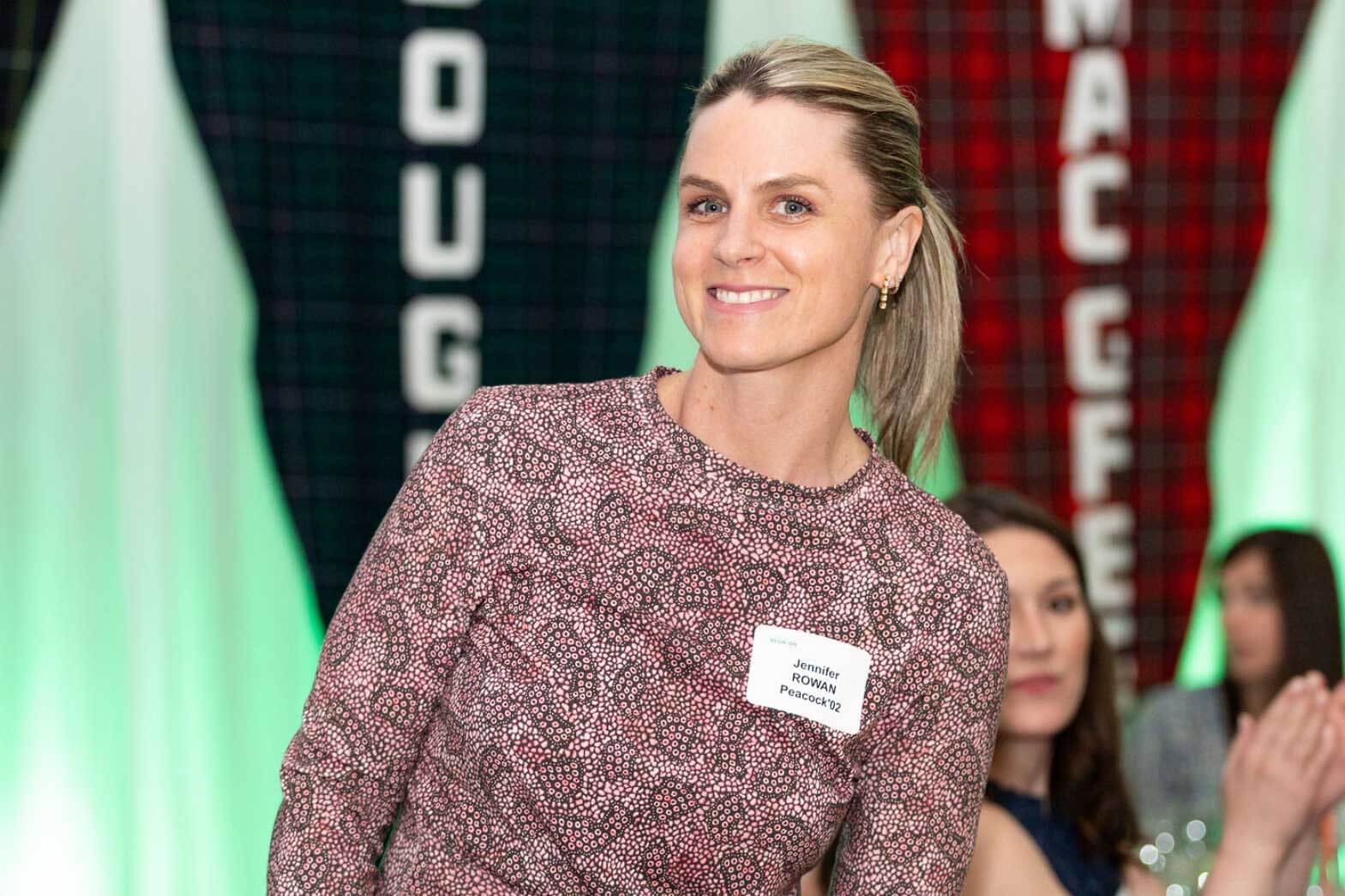 A woman with blonde hair in a ponytail, wearing a patterned dress and a name tag, smiles at the camera. She stands in front of colorful vertical banners and people seated at tables in the background.