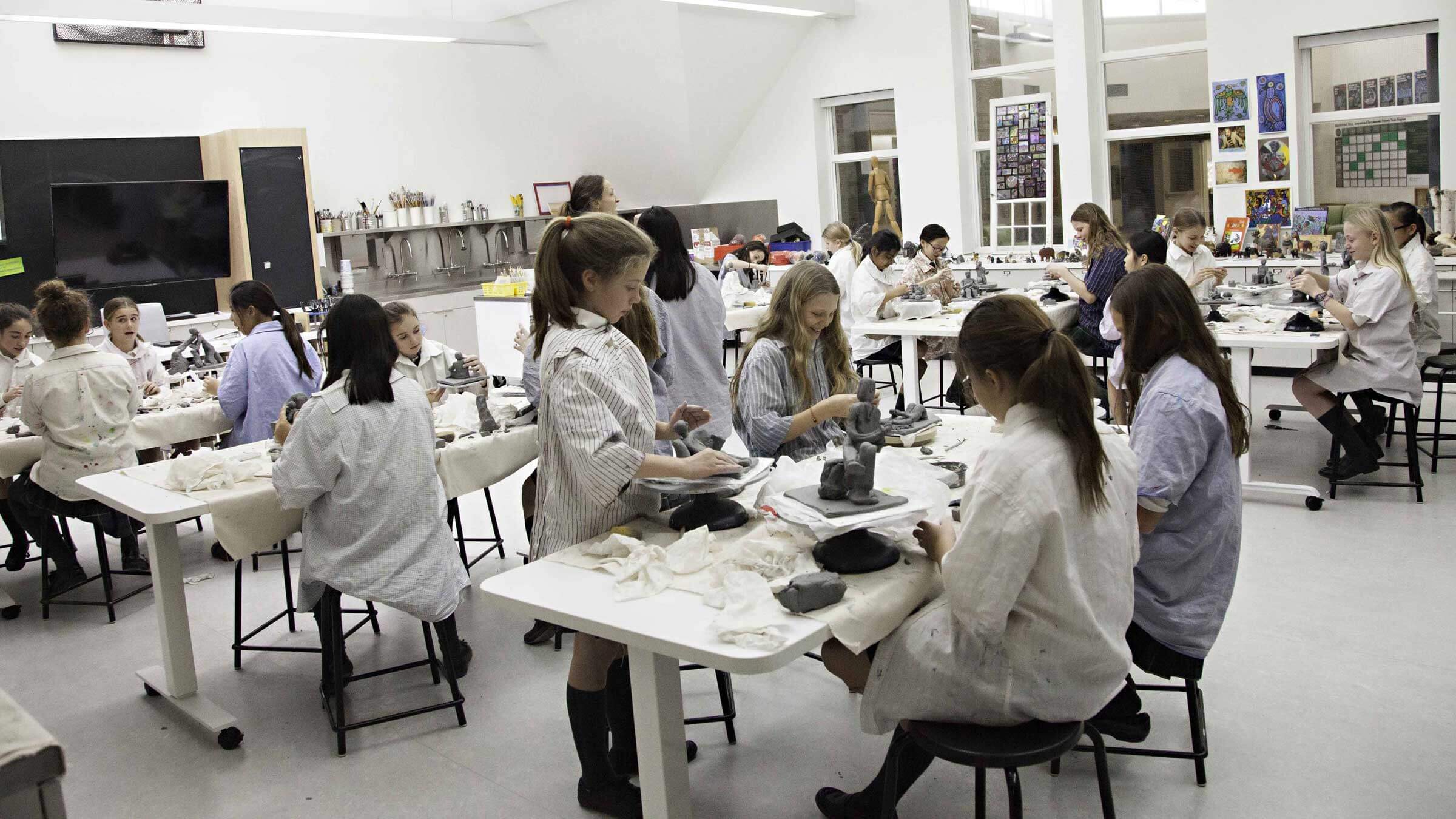 A classroom filled with students wearing smocks, sitting at tables and working with clay on sculpture projects. The room is bright, with large windows and various art supplies and artwork displayed on the walls.