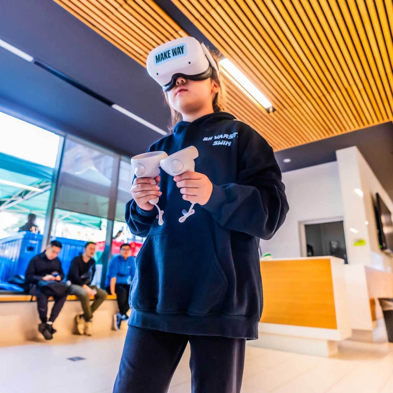 A child wearing a VR headset and holding controllers stands indoors, focused on a virtual experience. Several people sit in the background near a reception area with modern decor and a wooden ceiling.