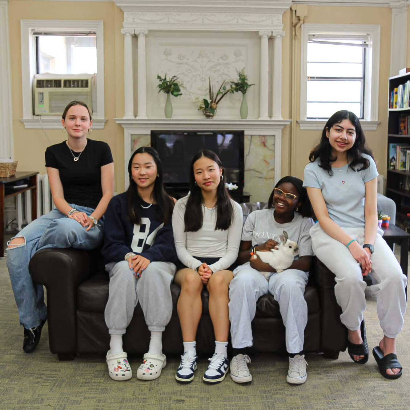 Five young women sit closely together on a brown couch in a cozy room; one holds a white bunny. They all smile at the camera, with bookshelves and a white fireplace in the background.