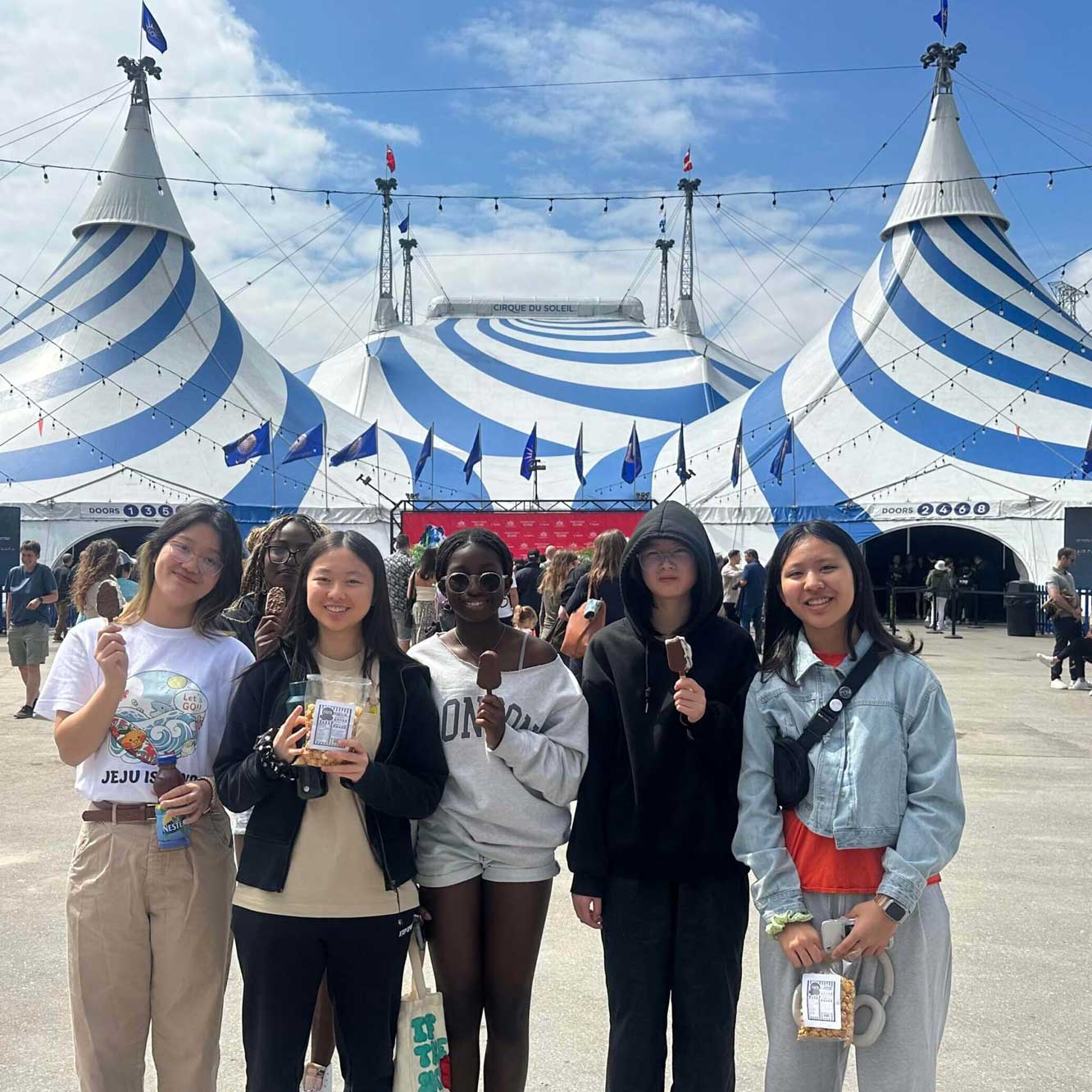 Five young people stand smiling and holding ice creams in front of a large blue and white circus tent, with a crowd and string lights in the background under a partly cloudy sky.