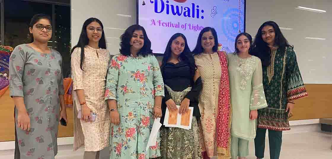 Seven women wearing traditional Indian attire stand together and smile in front of a screen displaying "Diwali: A Festival of Lights" at a brightly lit indoor event.