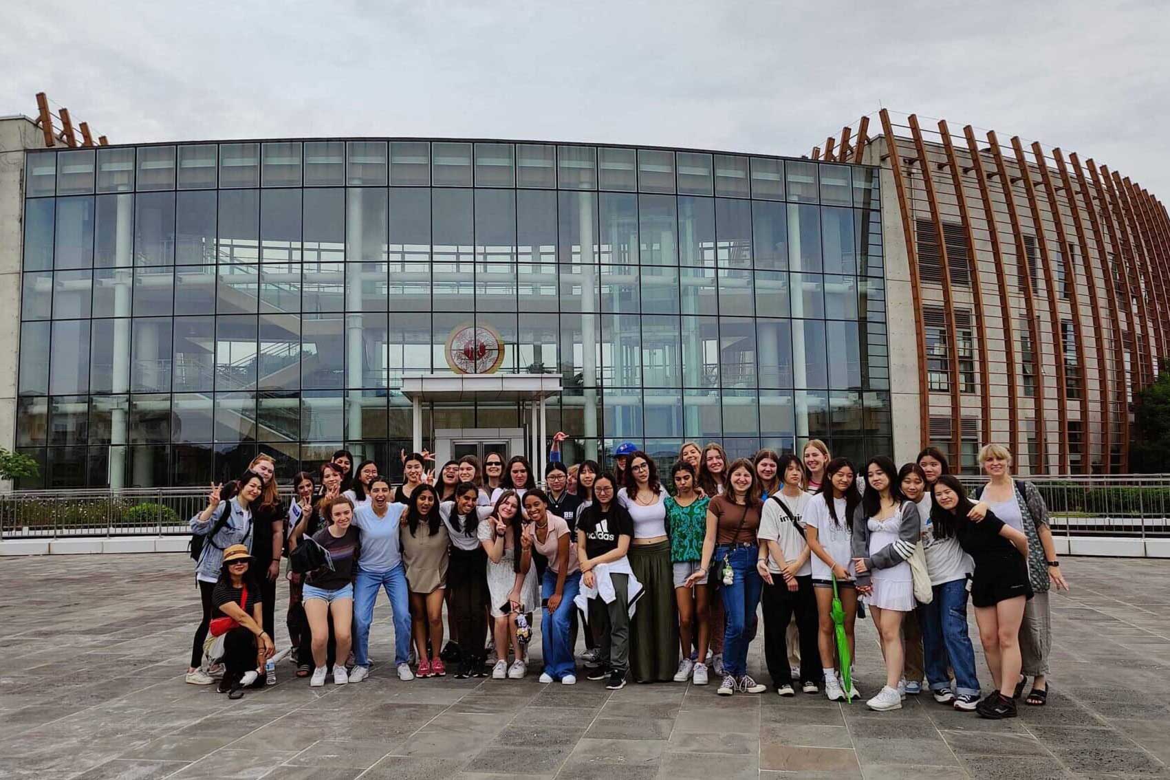 A large group of people poses for a photo in front of a modern glass building with wooden accents on a cloudy day. Most are smiling and casually dressed, standing and kneeling on an open plaza.