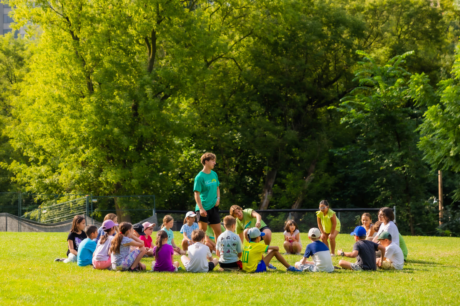 A group of children sit in a circle on a grassy field, listening to an adult standing in the center. Trees and greenery fill the background on a sunny day.