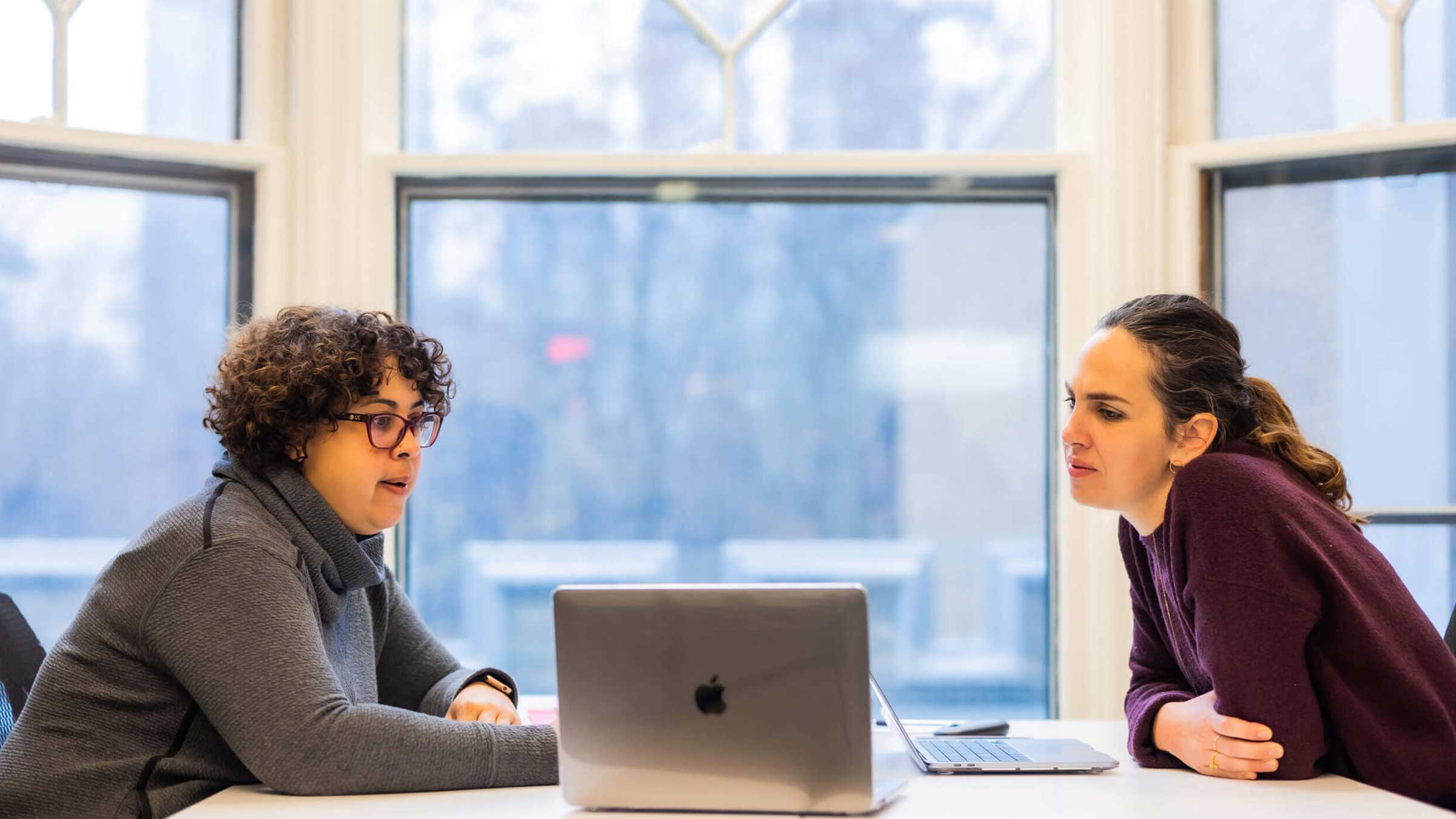 Two women sit at a table with a laptop between them, having a conversation in a bright room with large windows in the background.