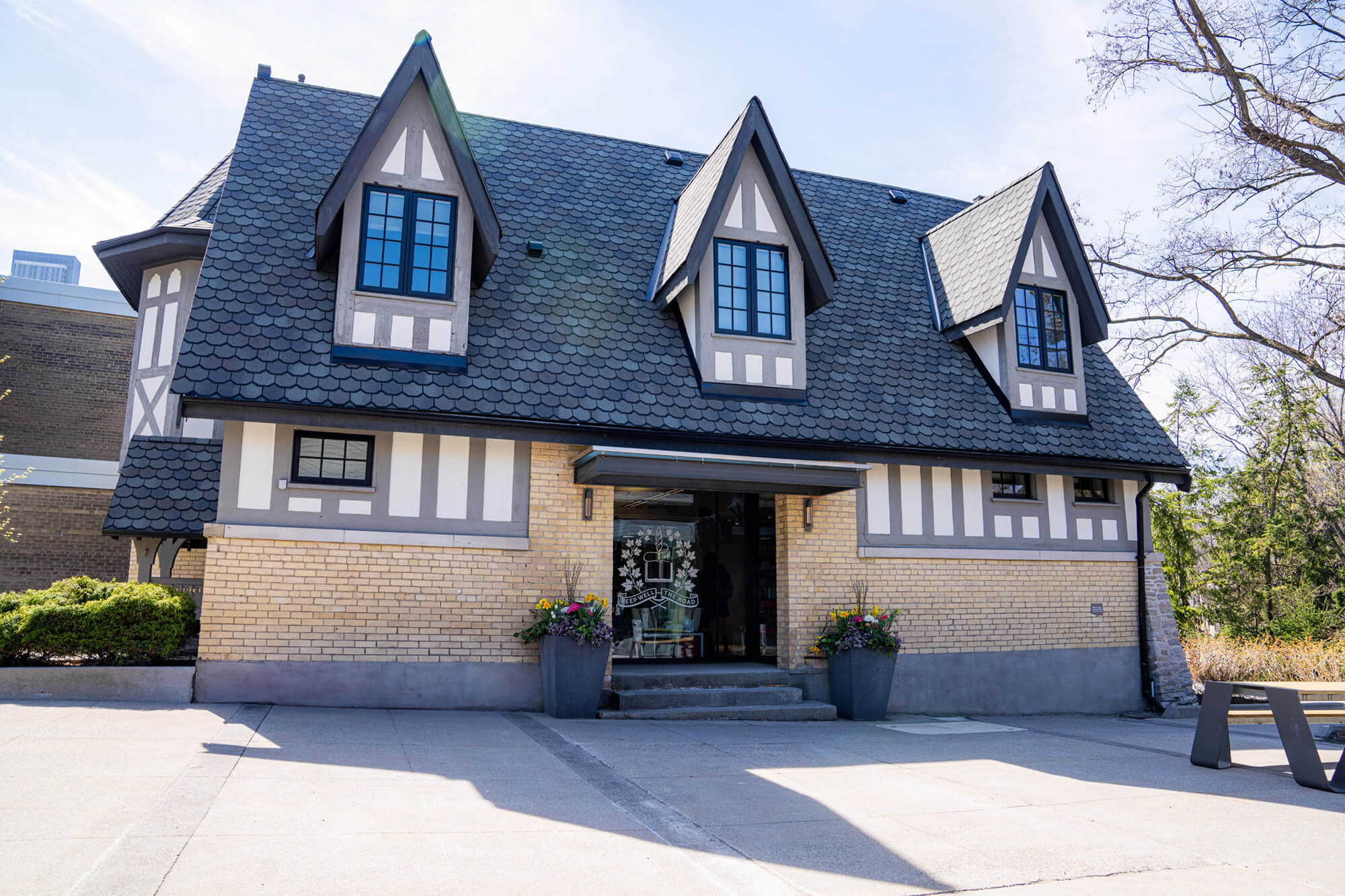 A two-story Tudor-style building with steep gabled roofs, black-and-white timbering, and tan brick walls. There are large planters with flowers by the entrance and trees in the background.