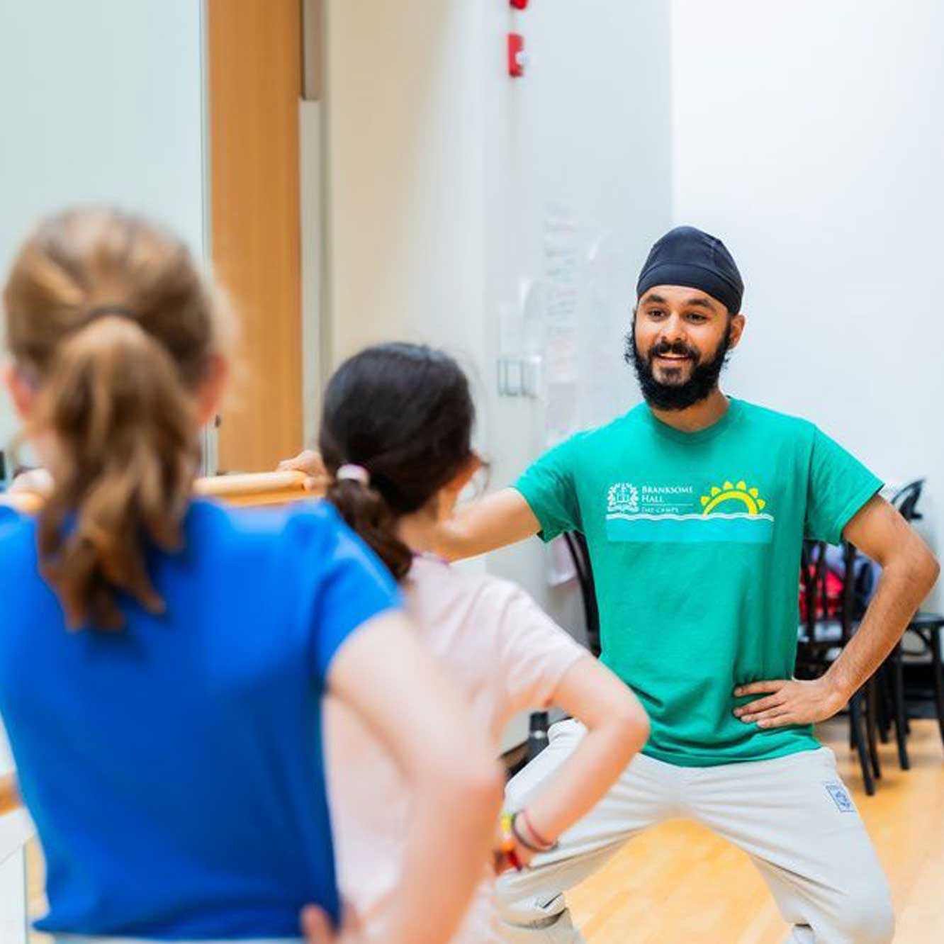 A man in a green t-shirt instructs two children in an exercise or dance class, standing with hands on hips and smiling, in a bright room with light-colored walls and wooden floors.