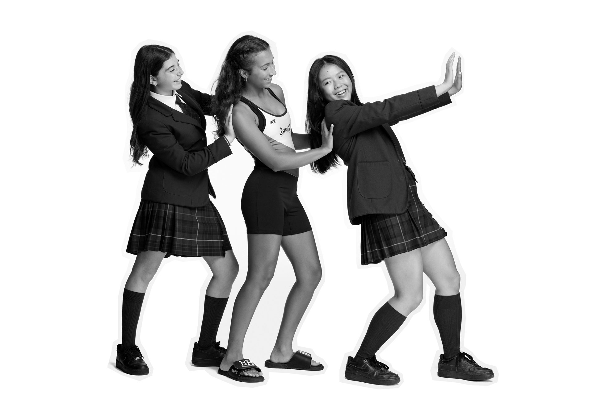 Three teenage girls stand in a line, smiling and playing. One wears a swimsuit, the other two wear school uniforms with skirts and blazers. They pose as if dancing or moving together in a fun, lighthearted manner.