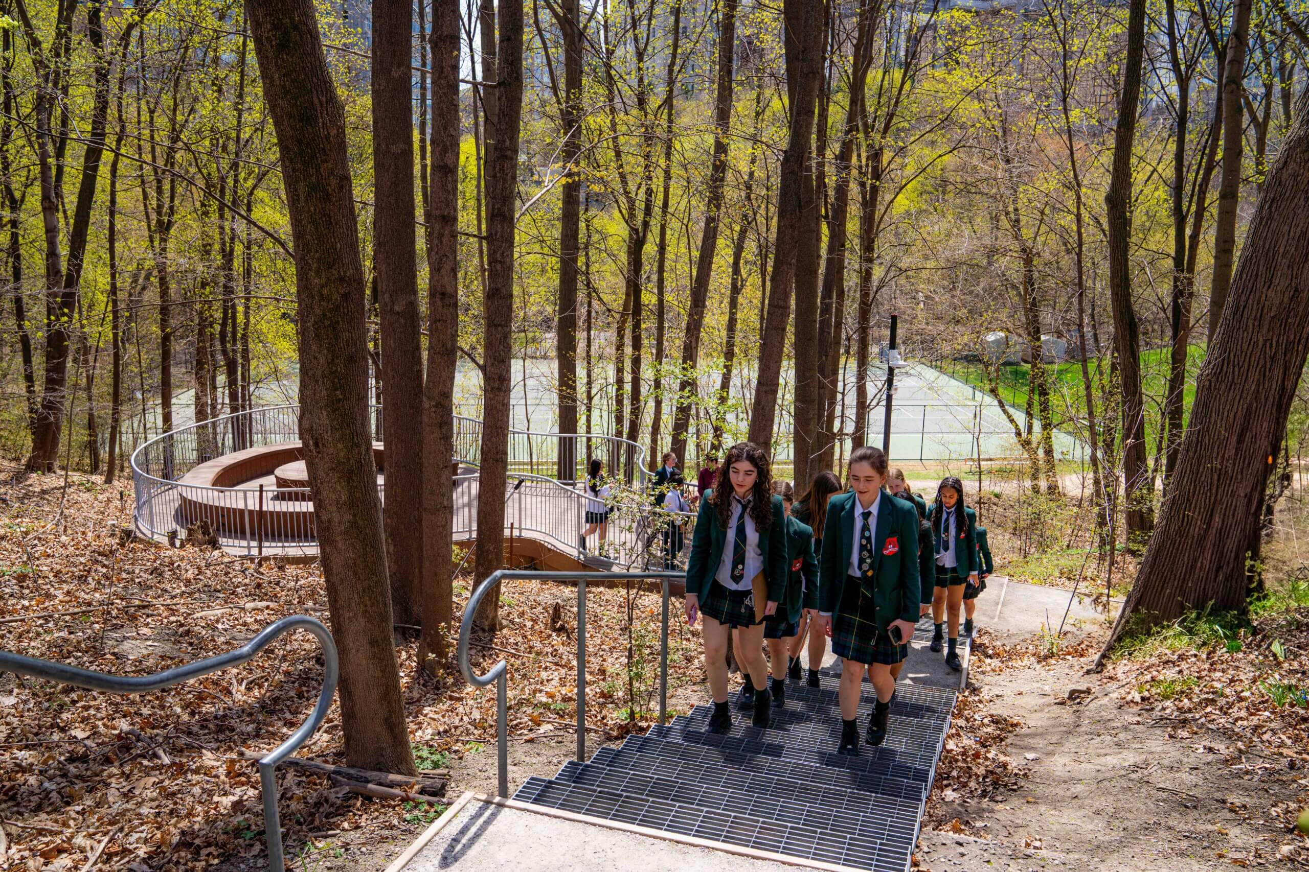 A group of students in school uniforms walk up metal stairs through a wooded area with bare trees and a greenhouse visible in the background.