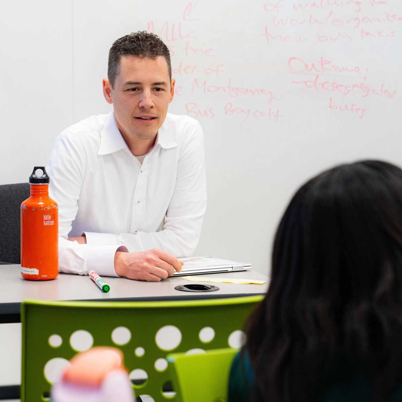 A man in a white shirt sits at a table with a notebook and orange water bottle, speaking to a person across from him in a classroom with notes written on a whiteboard in the background.